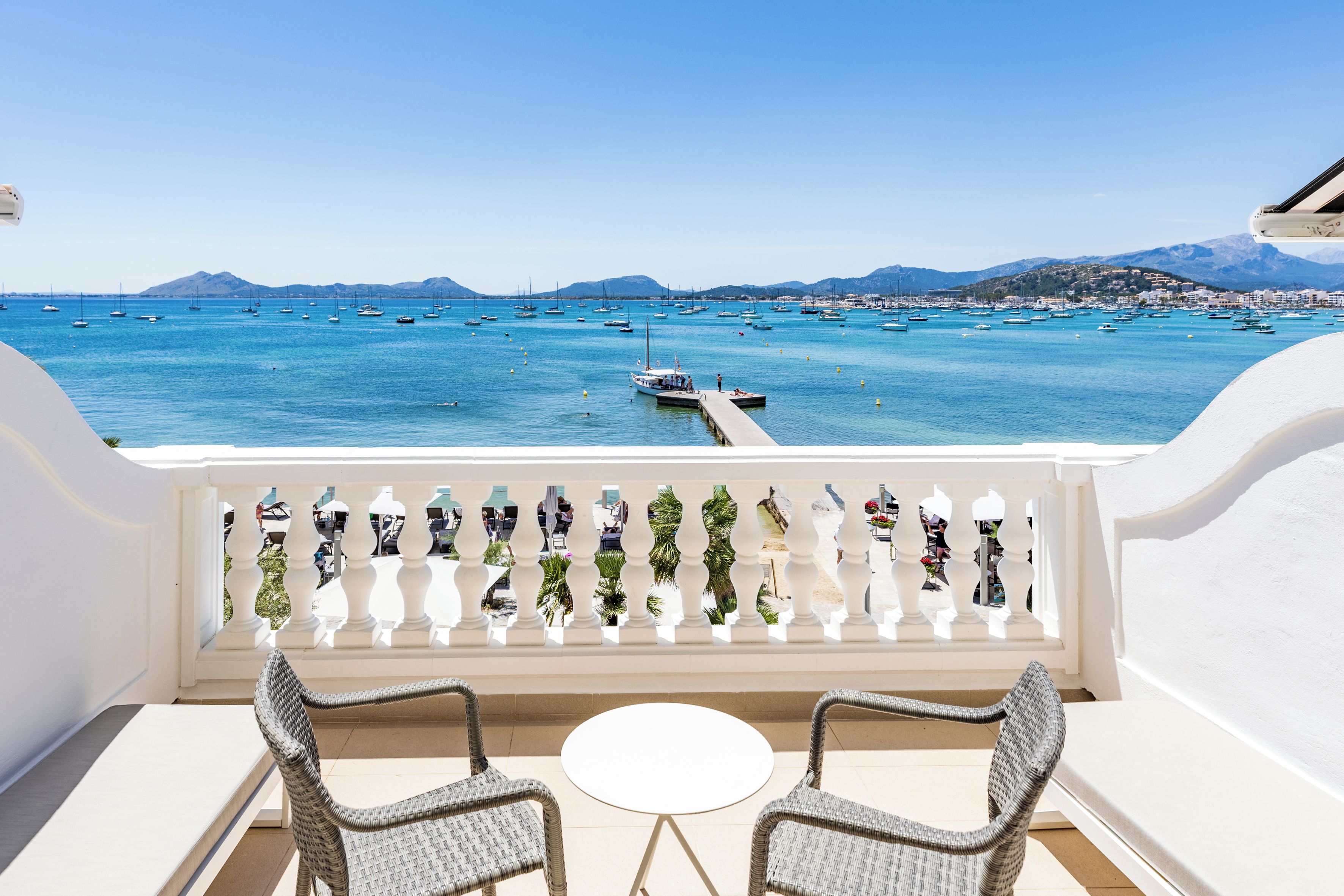 A balcony with two wicker chairs and a small round table, overlooking a marina with boats and a coastal town.