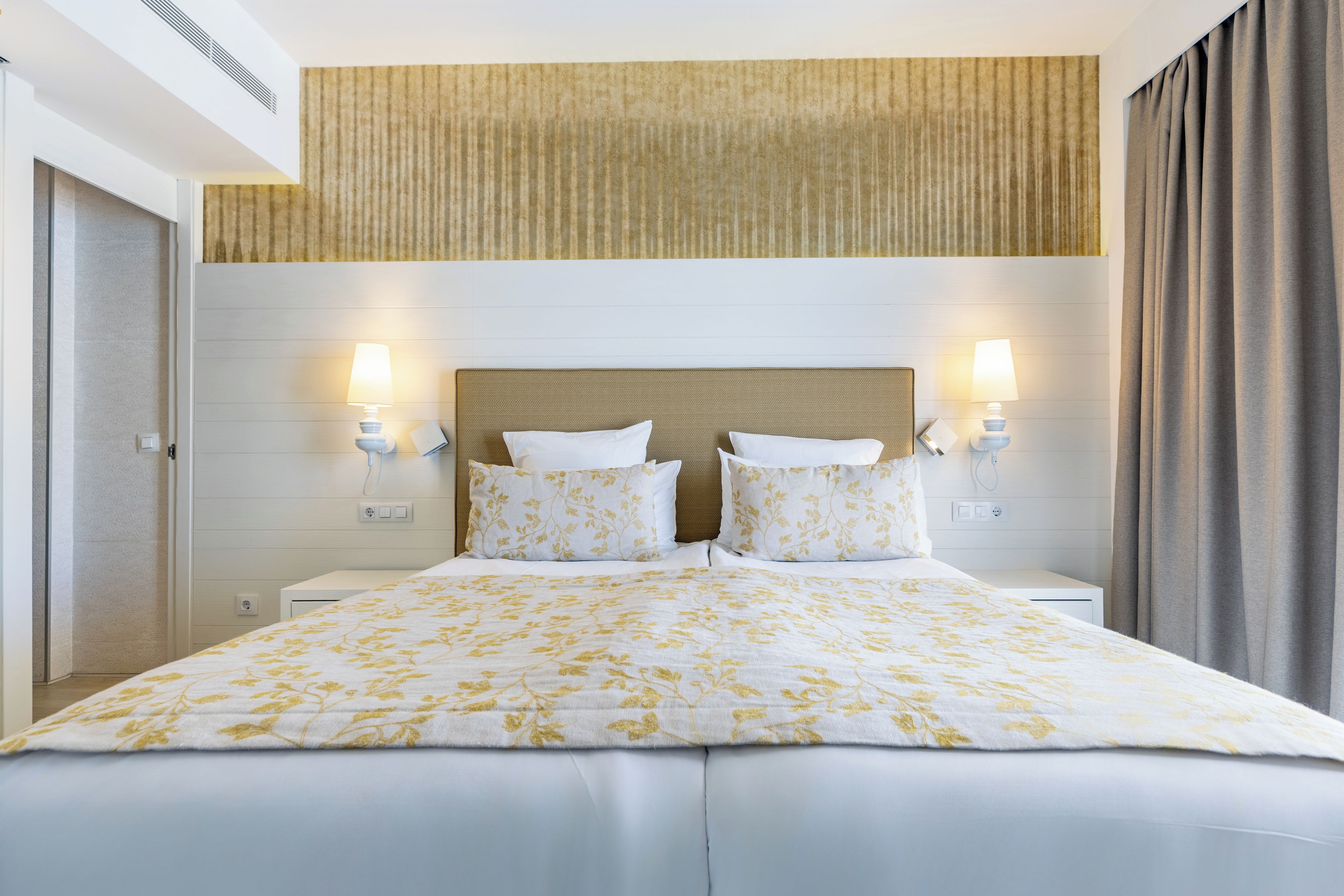Una habitación de hotel con una cama grande, dos lámparas de noche, un cabecero y una ventana con cortinas.