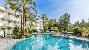 A swimming pool with lounge chairs and palm trees, surrounded by a multi-story building with balconies. - Hotel Illa D'Or (Pollenca)