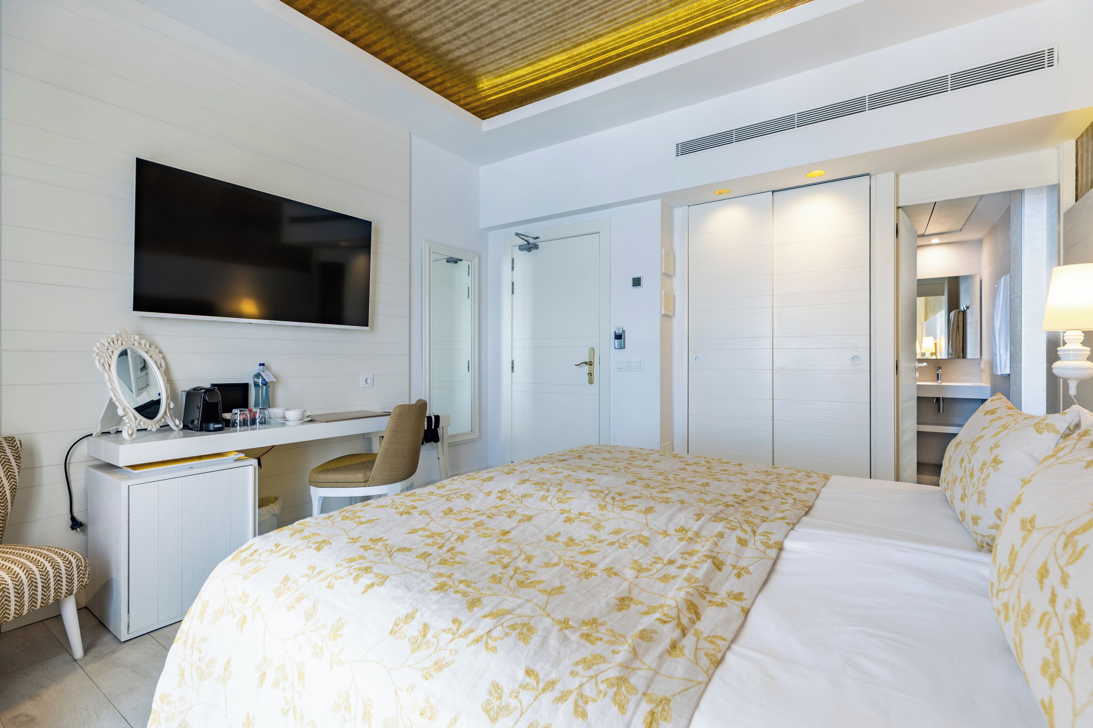Habitación de hotel con cama, escritorio, silla, televisor, armario y baño con espejo.