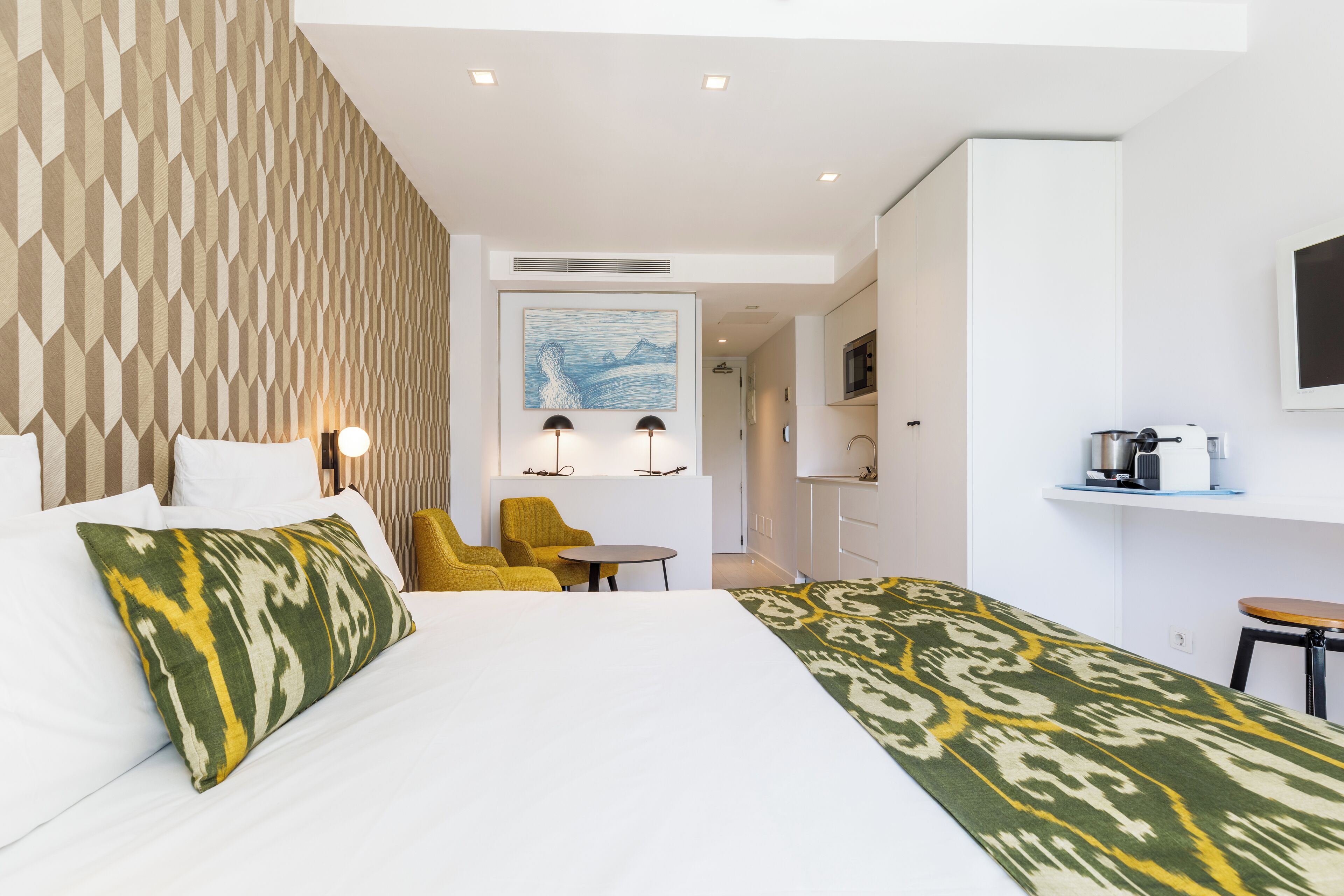 A hotel room with a bed, a small table, a chair, a TV, and a kitchenette.