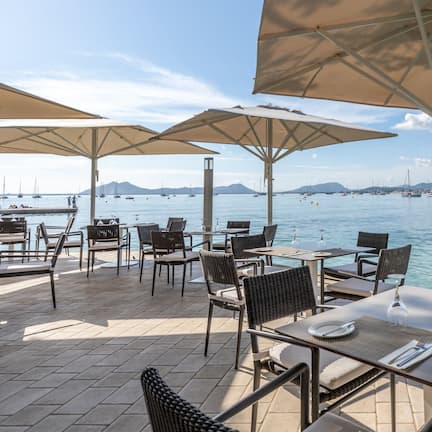 An outdoor dining area with tables and chairs under umbrellas, overlooking a body of water with boats and a distant cityscape.