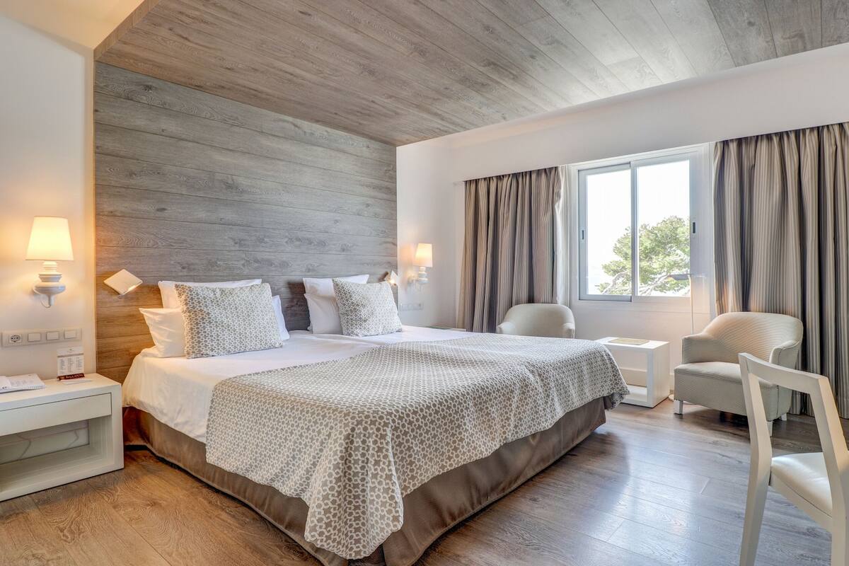 A modern hotel room with a wooden ceiling, a large bed with patterned bedding, a desk with a mirror, and a chair.