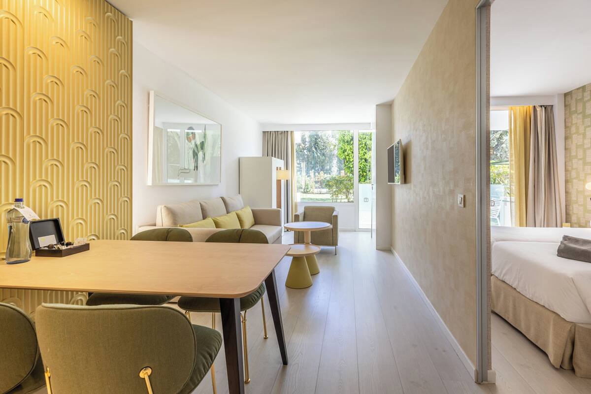 A hotel room with a bed, a sitting area with a sofa and armchairs, a dining table with chairs, and a large window providing a view of greenery outside.