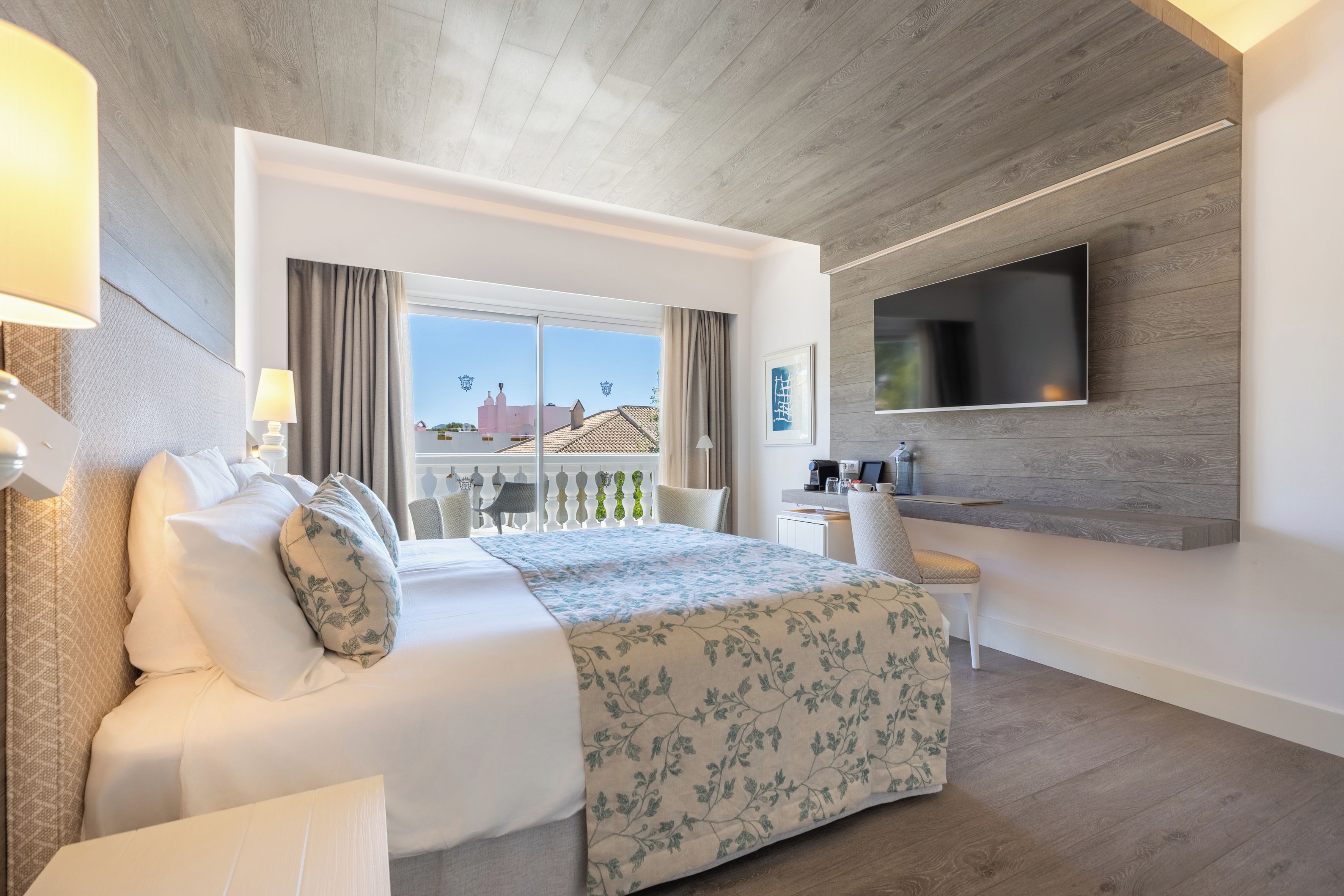 Ein Hotelzimmer mit Bett, Schreibtisch, Fernseher und einem Balkon mit Blick auf Gebäude.