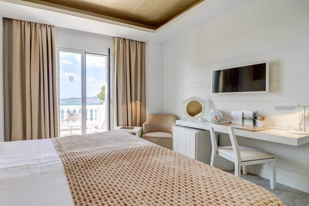 A hotel room with a bed, a desk, a chair, a TV, and a view of the sea.
