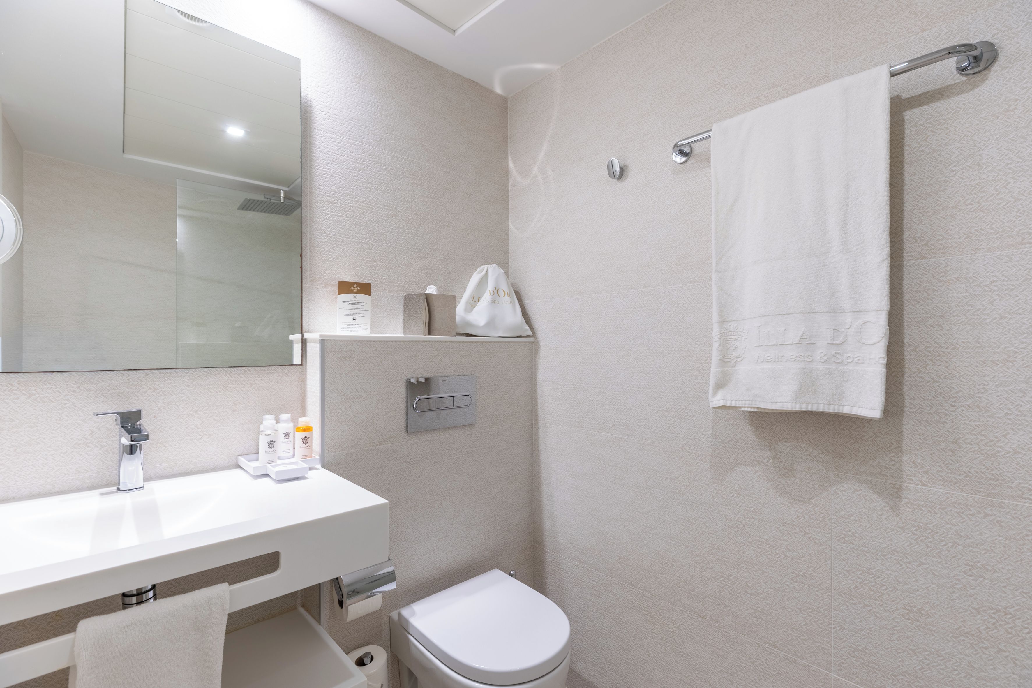 A modern bathroom with a white sink, mirror, and toilet. There is a towel rack with a white towel hanging, and toiletries are placed on the sink.