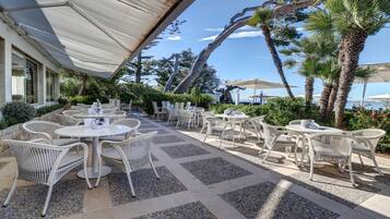 Une terrasse extérieure avec des tables rondes et des chaises en osier, entourée de palmiers et offrant une vue sur la mer.