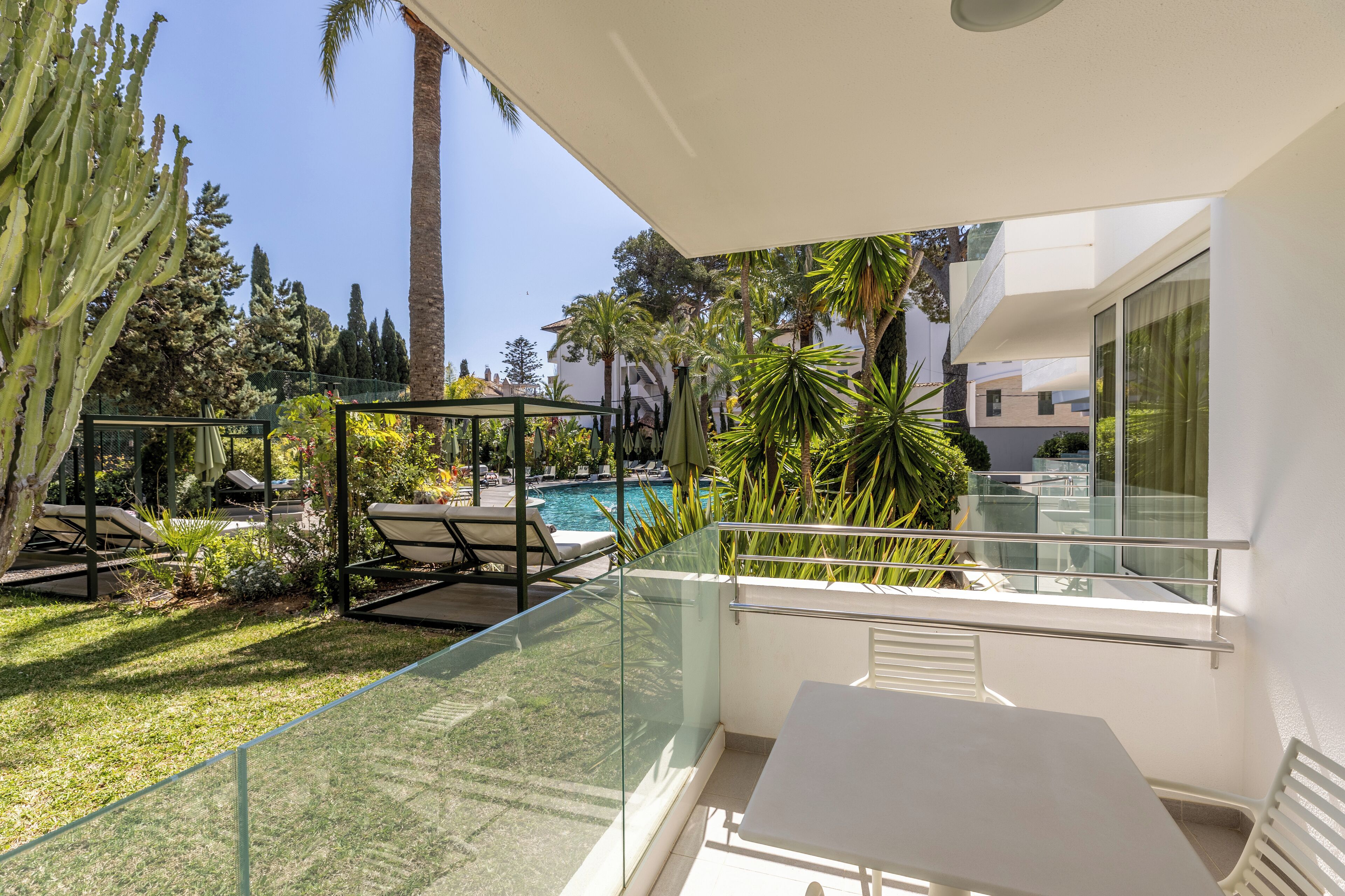 Un balcon moderne avec des garde-corps en verre donnant sur une piscine, entouré d’une végétation luxuriante et de palmiers.