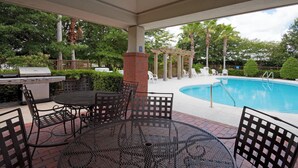 Eine Terrasse im Freien mit Grill, rundem Tisch und Stühlen, im Hintergrund ein Pool.