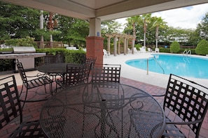 Un patio al aire libre con una parrilla, una mesa redonda y sillas, y una piscina al fondo.