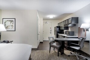Une chambre d’hôtel comprenant un lit, un bureau avec une chaise, un four à micro-ondes, un réfrigérateur et une petite table avec une lampe.