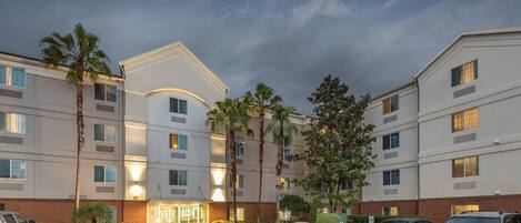 A multi-story hotel with a central entrance, parking area, and surrounding greenery.