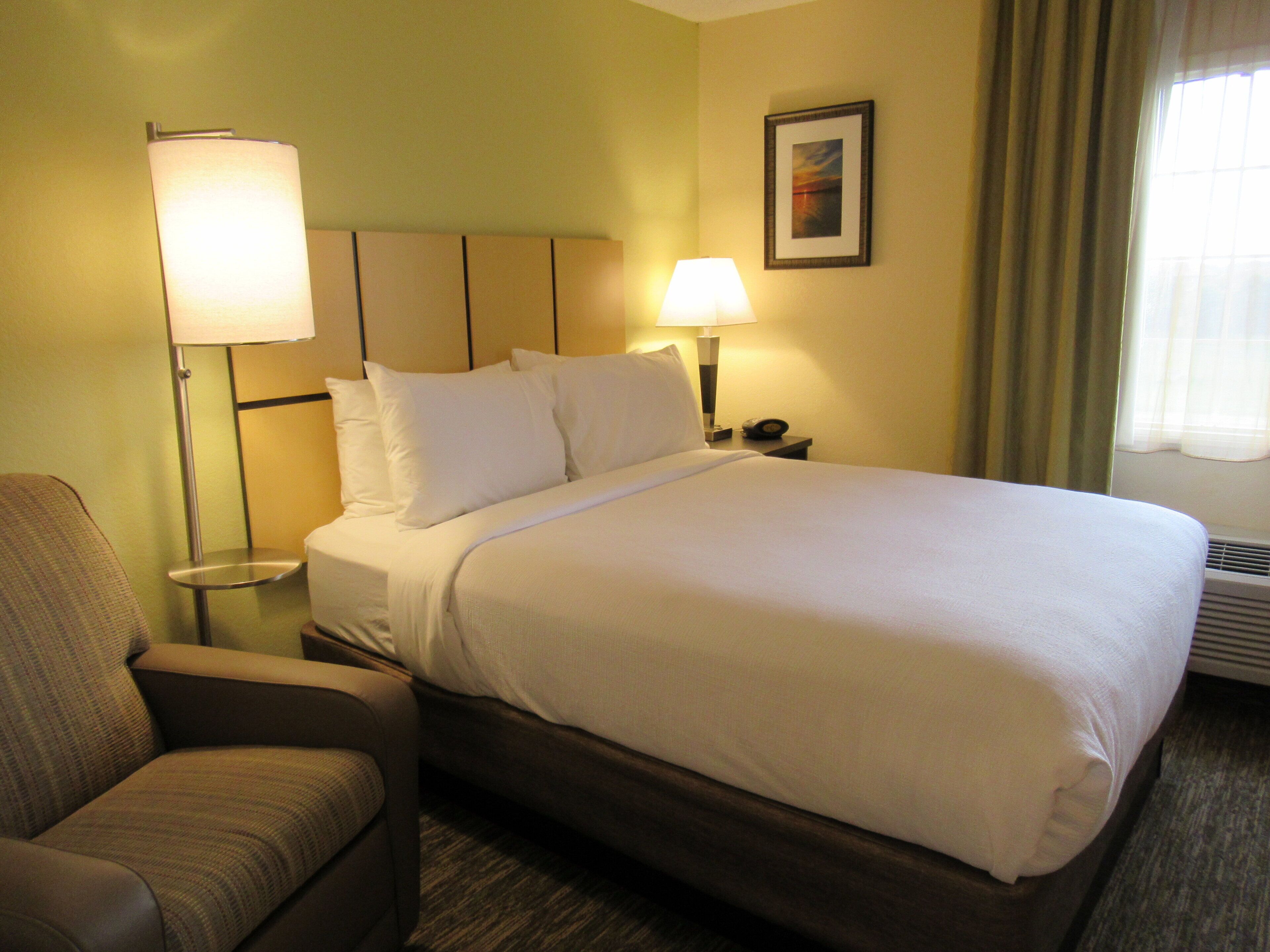 A hotel room with a bed, two bedside lamps, a chair, and a framed picture on the wall.