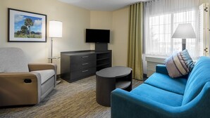 Hotelkamer met een beige muur, een lijst met foto, een flatscreen televisie, een blauwe bank, een klein rond tafeltje en een lamp.