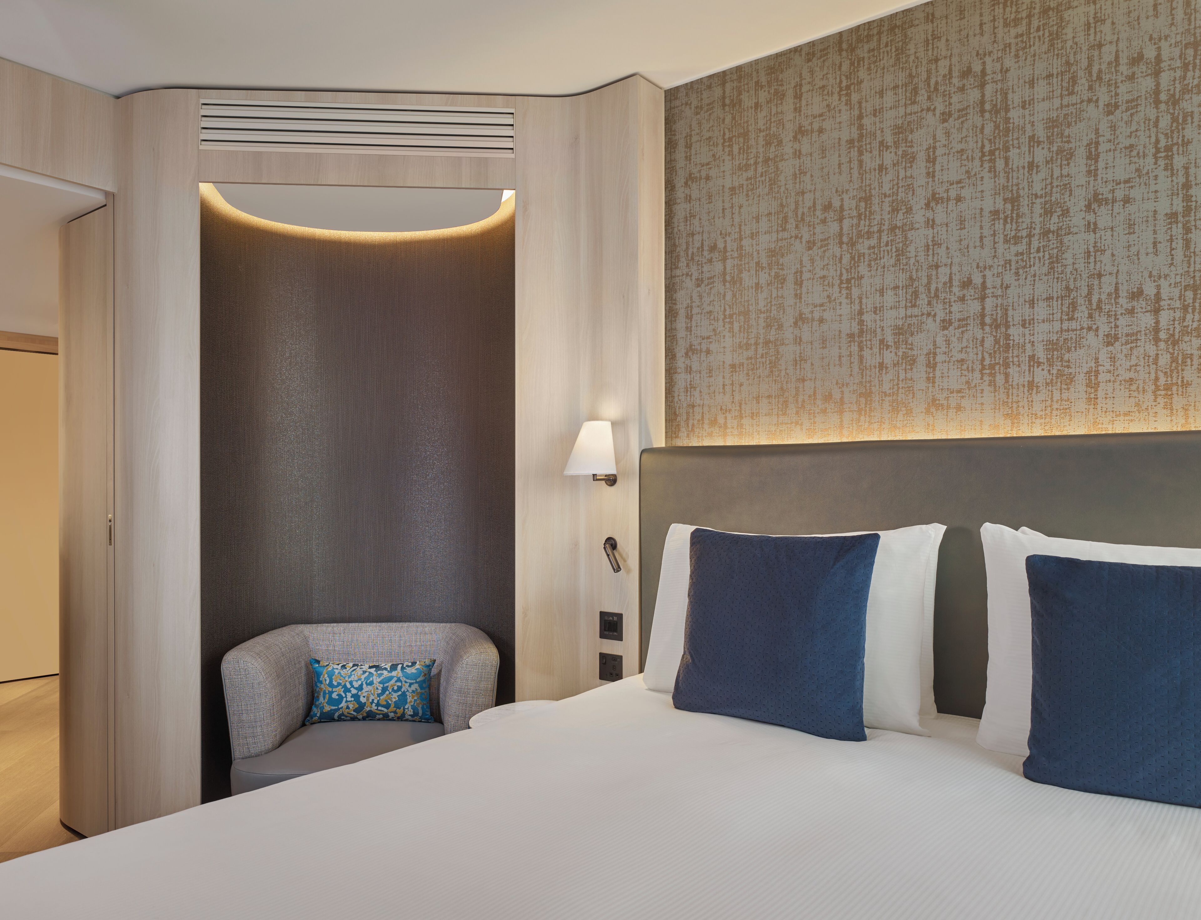 A hotel room with a bed, a seating area, a wall-mounted lamp, and a textured wall.