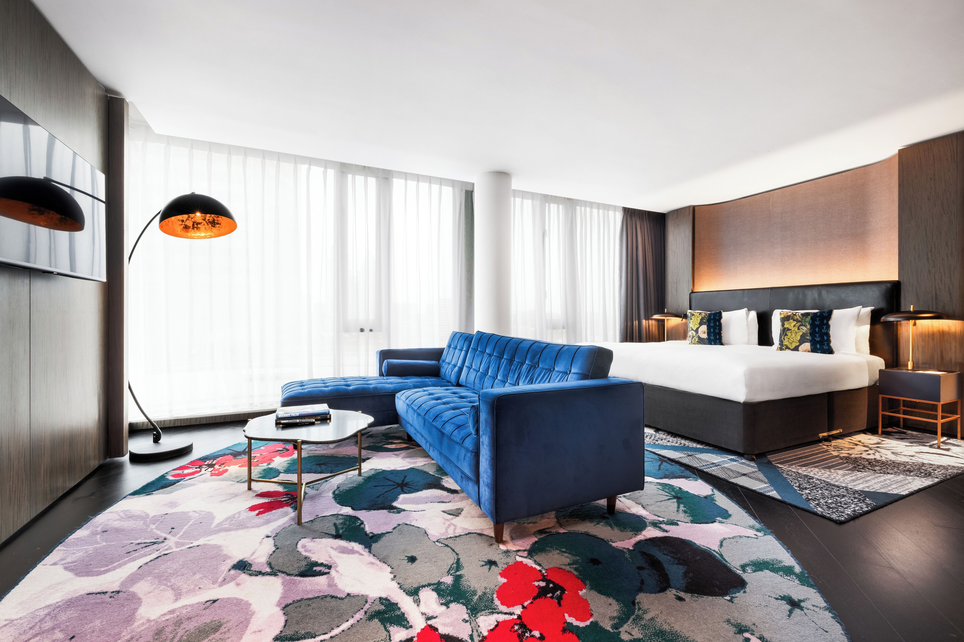 Ein modernes Hotelzimmer mit Ecksofa, einem Bett mit vielen Kissen, einem großen Fenster mit durchsichtigen Vorhängen und einer Stehlampe.