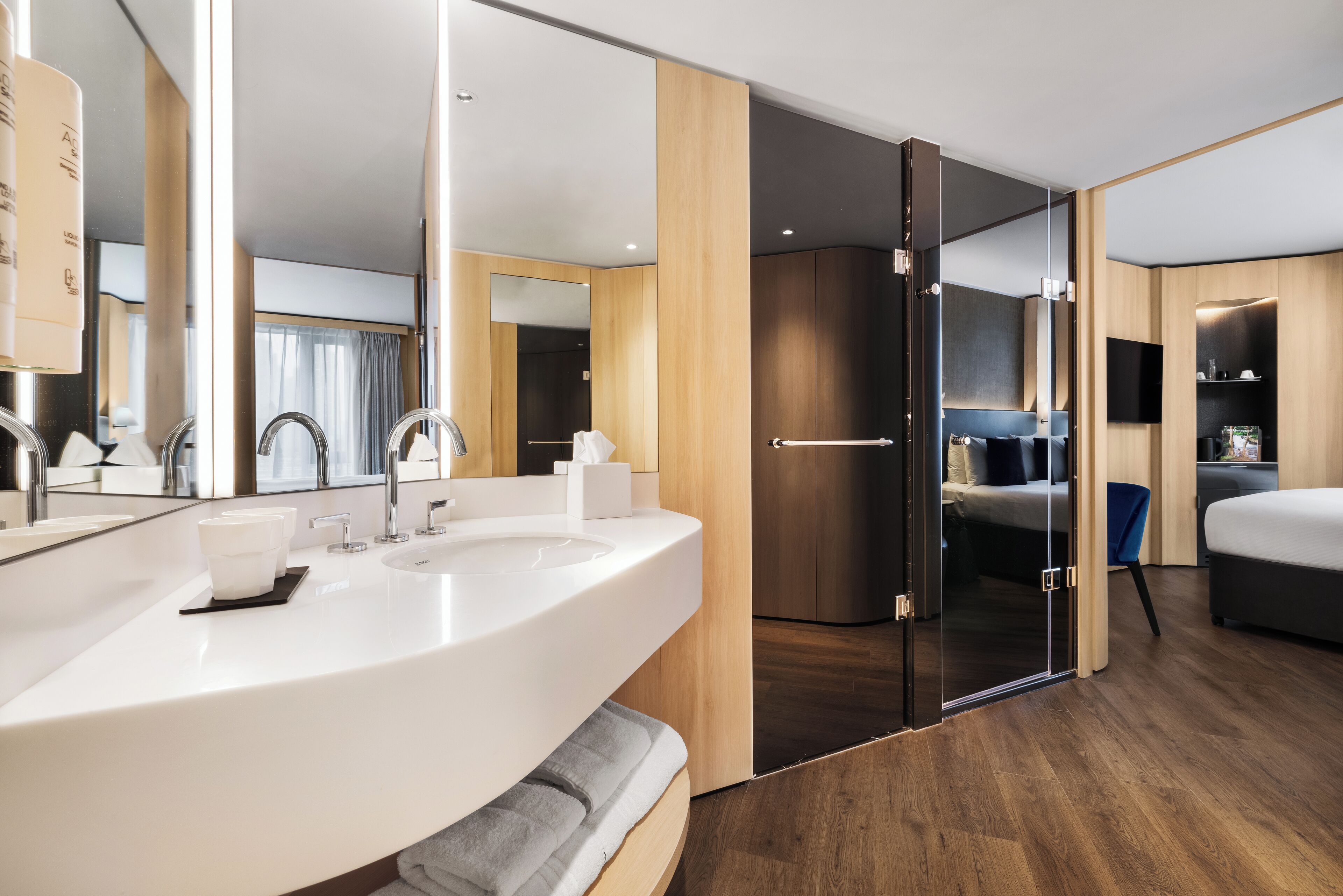 A modern hotel room with a large white sink, a mirror, a flat-screen TV, a bed, and a sliding door leading to a walk-in closet.