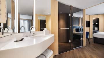 Ein modernes Hotelzimmer mit einem großen weißen Waschbecken, einem Spiegel, einem Flachbildfernseher, einem Bett und einer Schiebetür zu einem begehbaren Kleiderschrank.