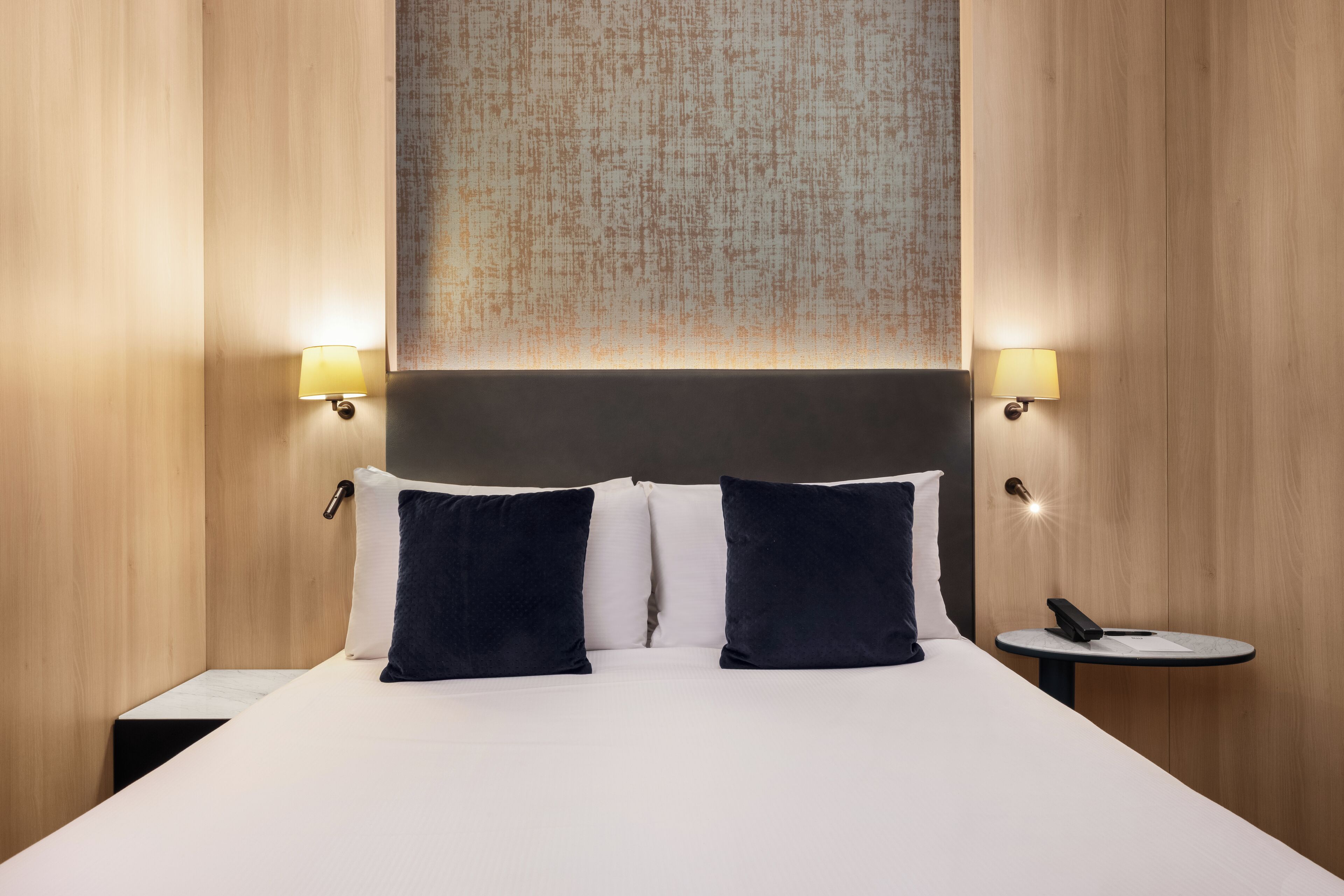 A hotel room with a large bed, two bedside lamps, a small table with a phone, and a textured wall hanging above the bed.
