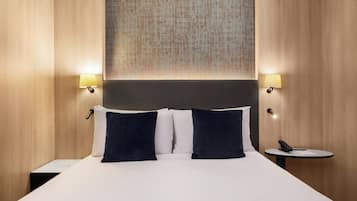 A hotel room with a large bed, two bedside lamps, a small table with a phone, and a textured wall hanging above the bed.