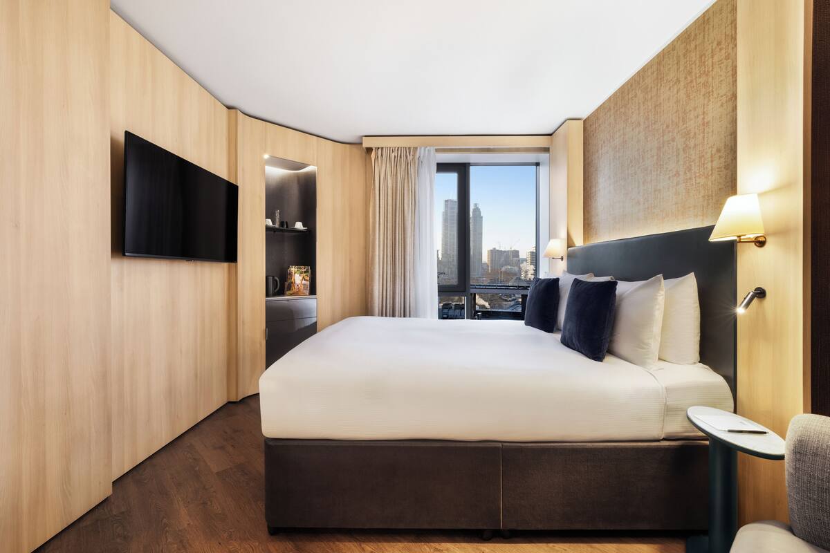 A modern hotel room with a large bed, a flat-screen TV, a small table, and a view of the city through a large window.