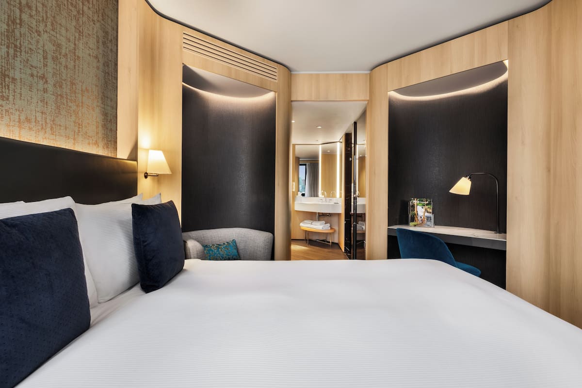 A modern hotel room with a large bed, a desk, and a bathroom visible through an open door.