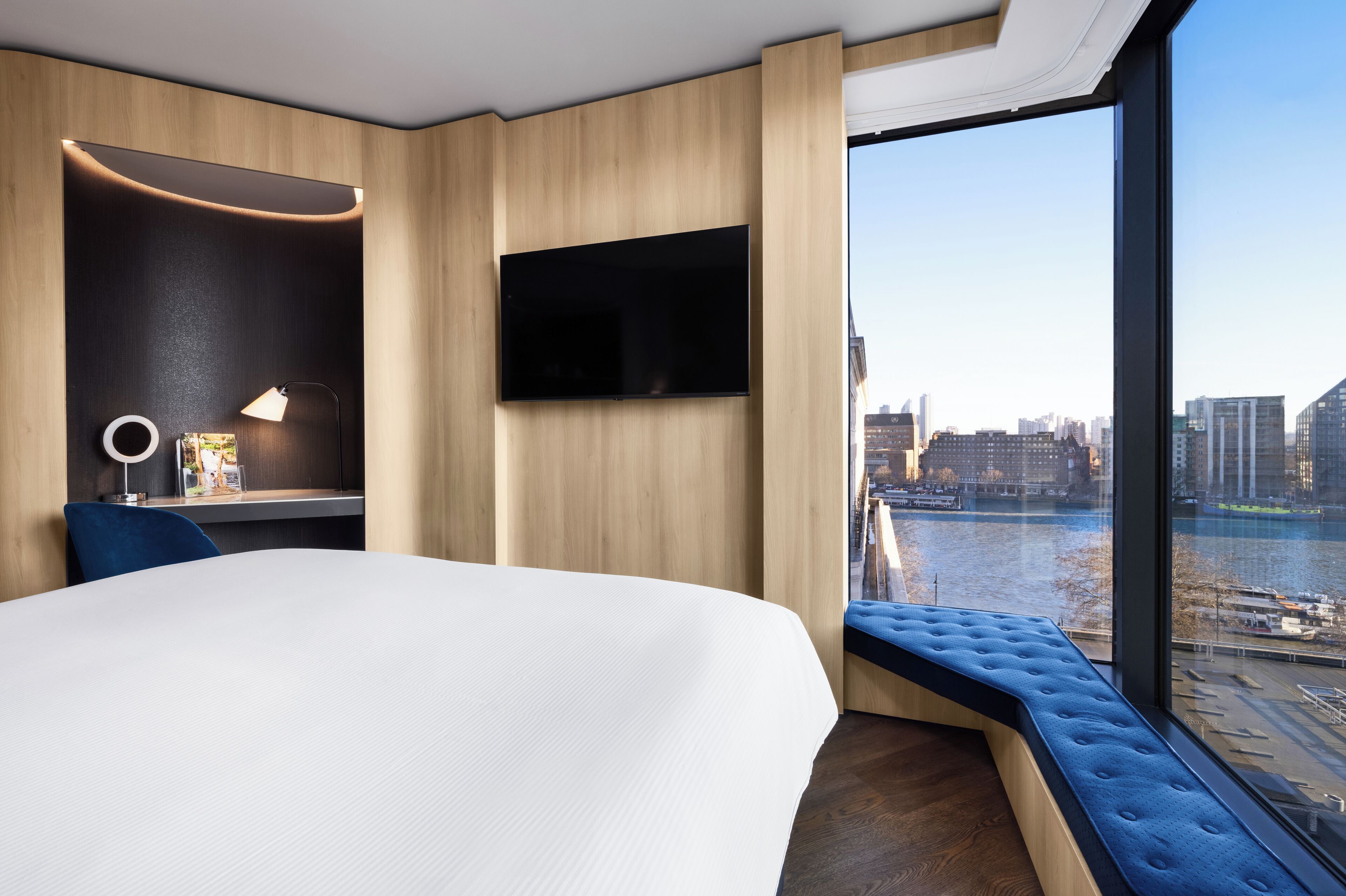 A modern hotel room with a large bed, a desk with a lamp, a flat-screen TV, and a view of a cityscape and river.