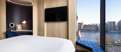 Una habitación de hotel moderna con una cama grande, un escritorio con lámpara, un televisor de pantalla plana y vistas a la ciudad y al río.