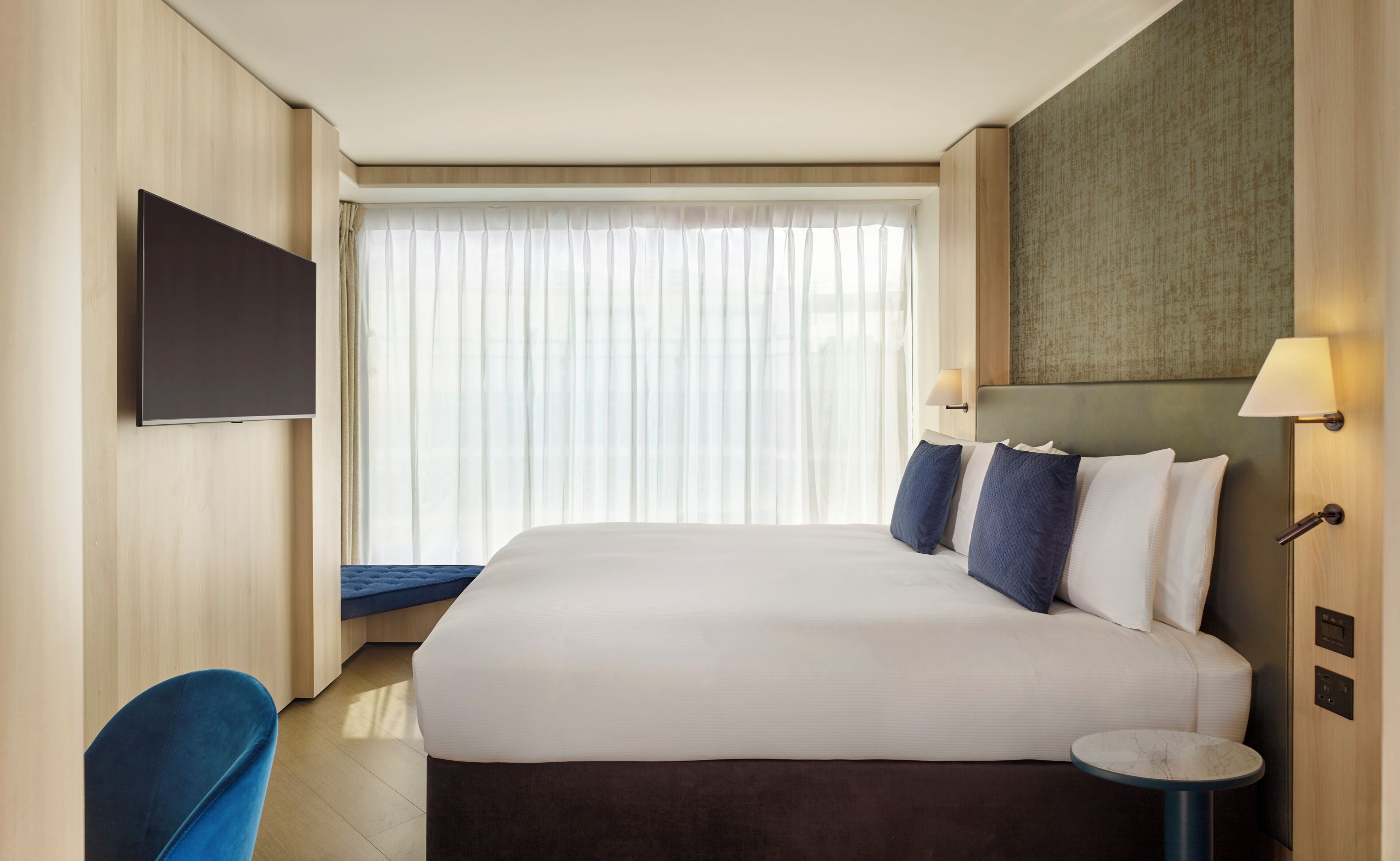 Ein Hotelzimmer mit einem großen Bett, einem wandmontierten Flachbildfernseher, einem Nachttisch und einem Fenster mit transparenten Vorhängen.