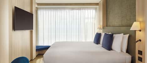 A hotel room with a large bed, a flat-screen TV mounted on the wall, a bedside table, and a window with sheer curtains.