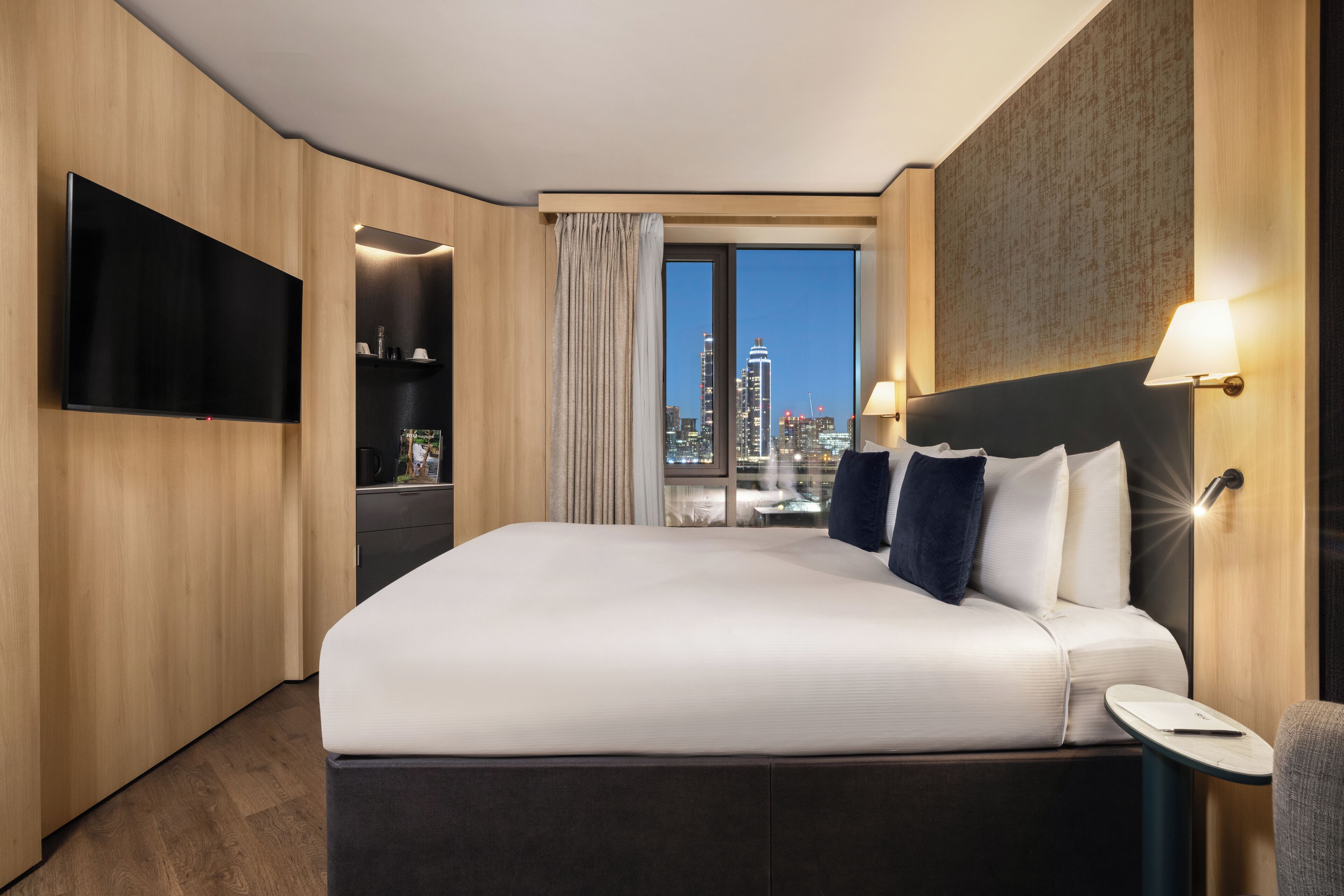 A modern hotel room with a large bed, a flat-screen TV, a nightstand, and a view of the city skyline.