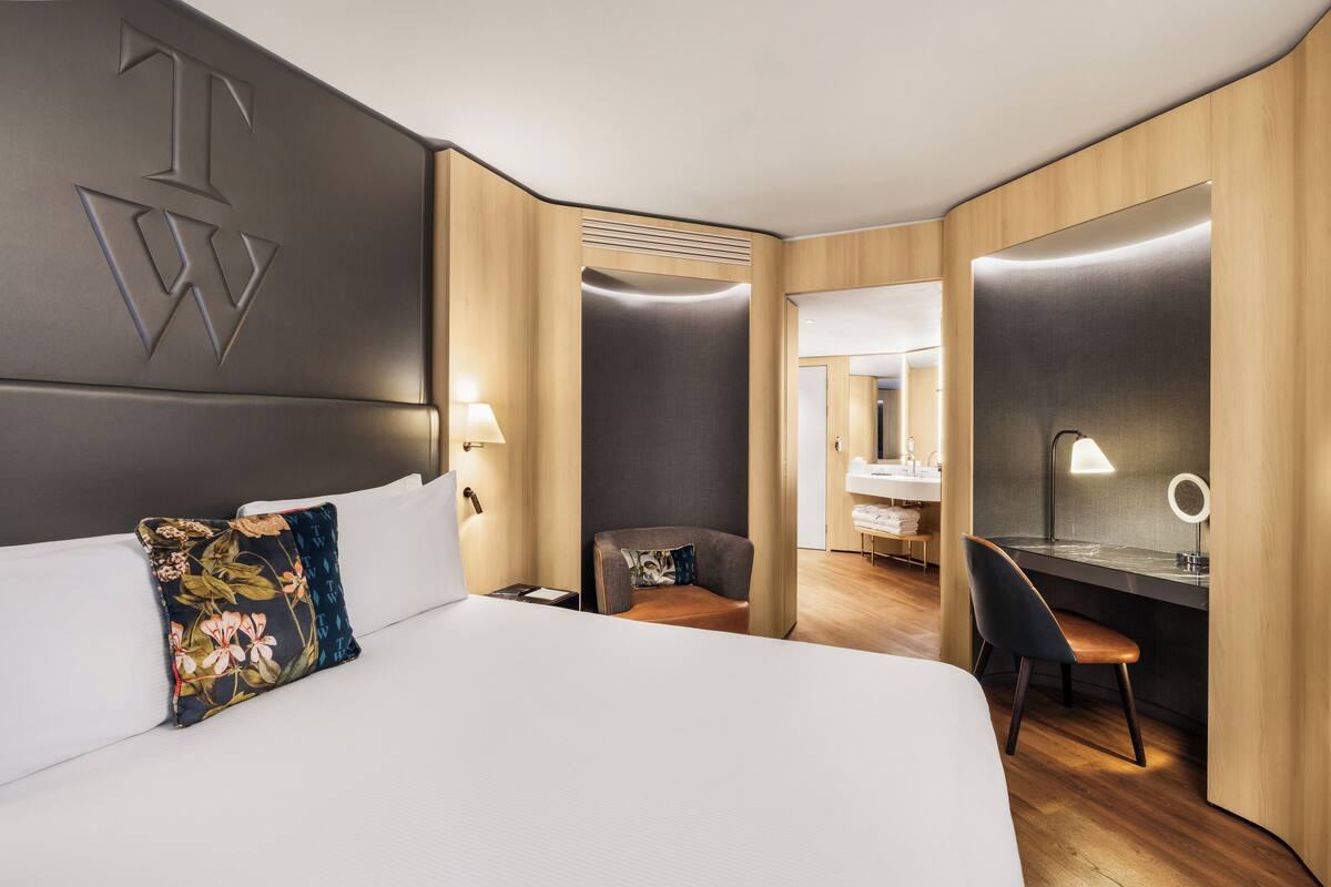 A hotel room with a large bed, a desk with a chair, a mirror, and a bathroom visible through an open door.