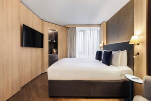 Ein modernes Hotelzimmer mit einem großen Bett, einem wandmontierten Flachbildfernseher, einem Nachttisch mit Lampe und Blick ins Freie durch durchsichtige Vorhänge.