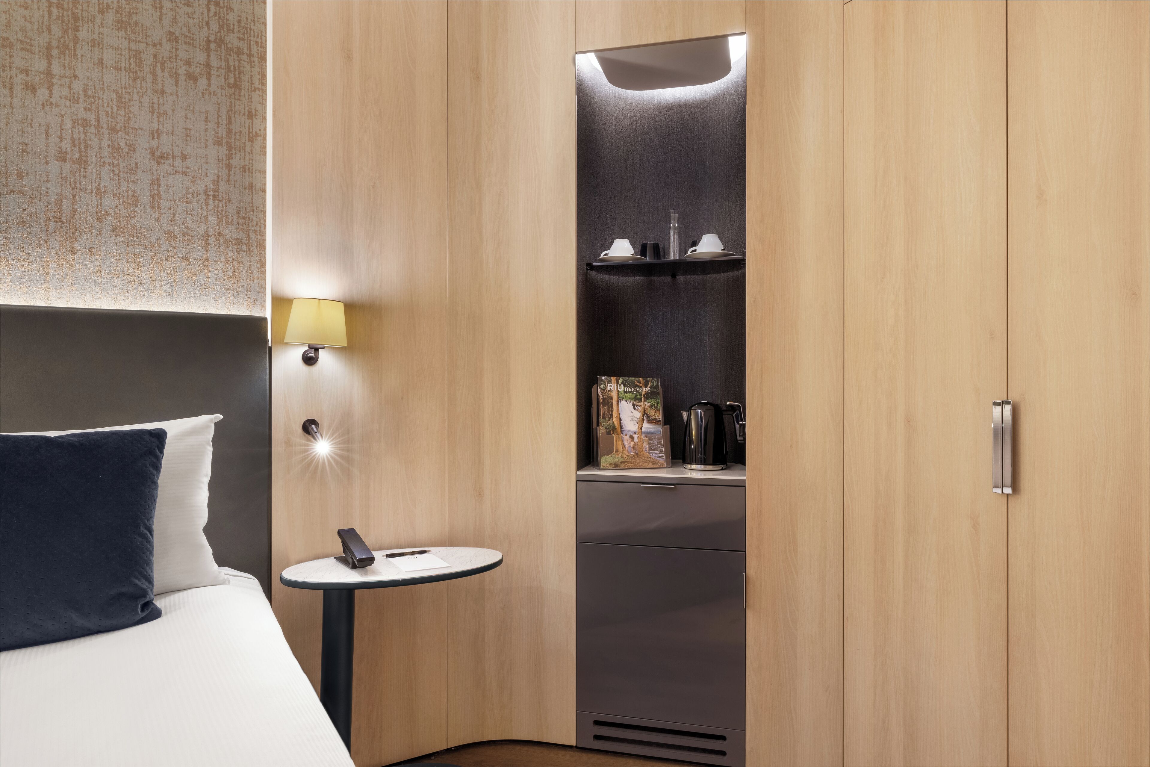 Een moderne hotelkamer met een bed, nachtkastje, lamp en een ingebouwde minibar.