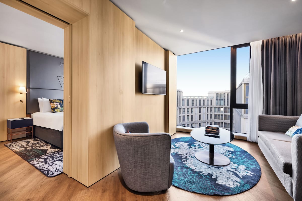 A modern hotel room with a bed, a gray armchair, a round coffee table, and a large window offering a city view.