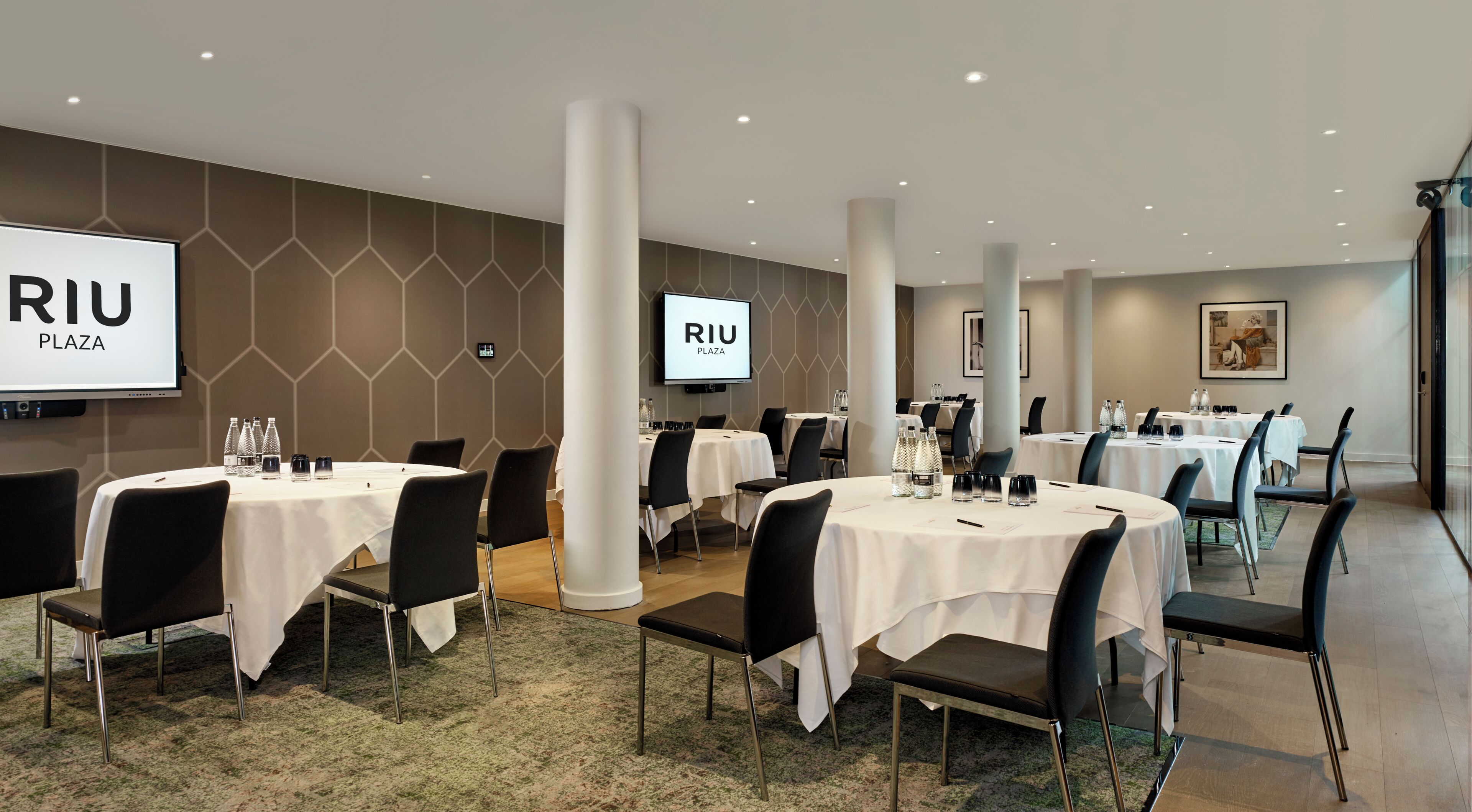 A conference room with multiple tables, chairs, and a large screen displaying 'RIU PLAZA'.