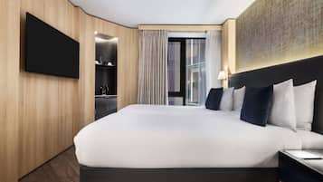 A modern hotel room with a large bed, a flat-screen TV, a desk with a lamp, and a view of the city through the window.