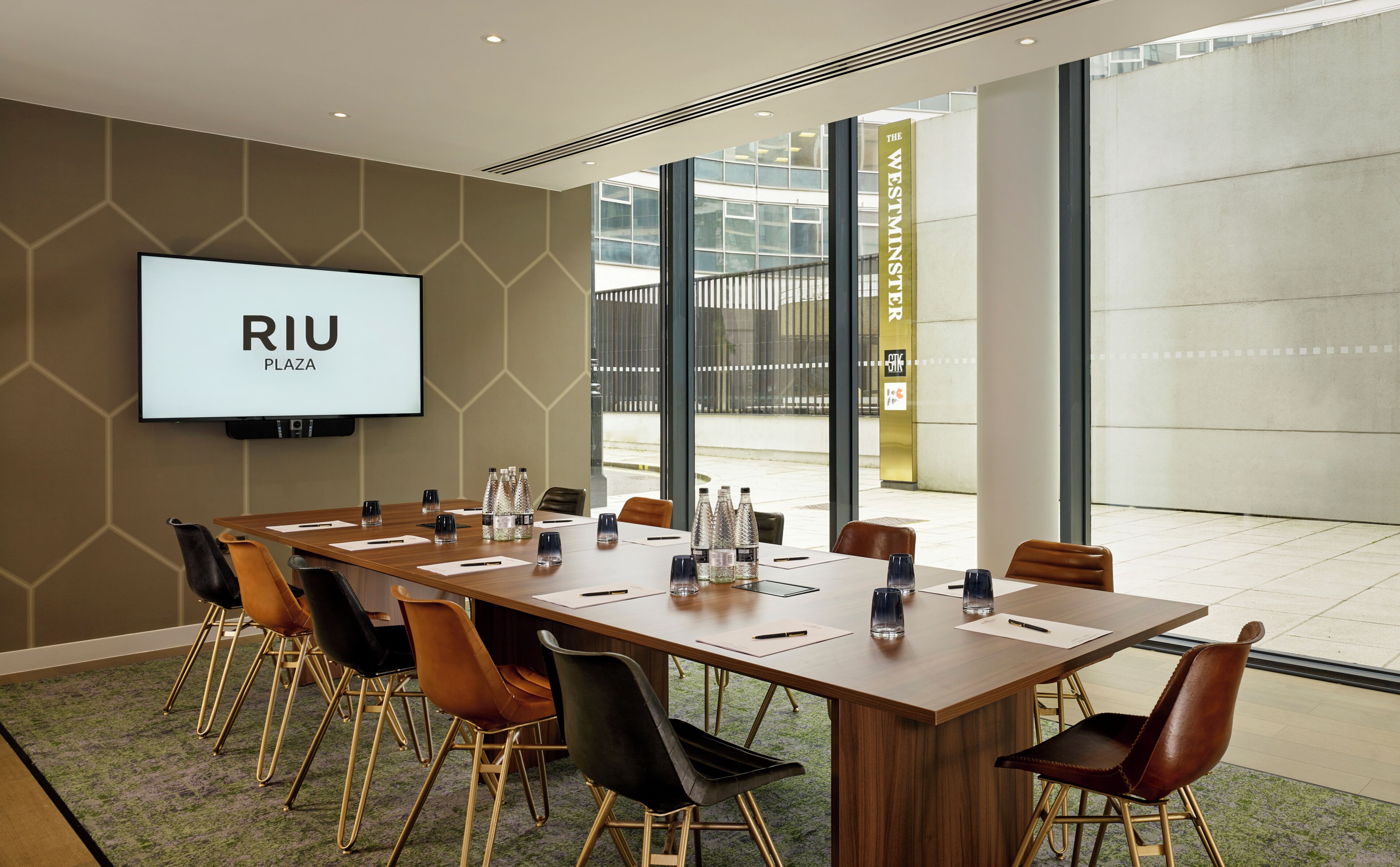 A conference room with a long table, chairs, a TV screen displaying 'RIU PLAZA', and a view of the outside.