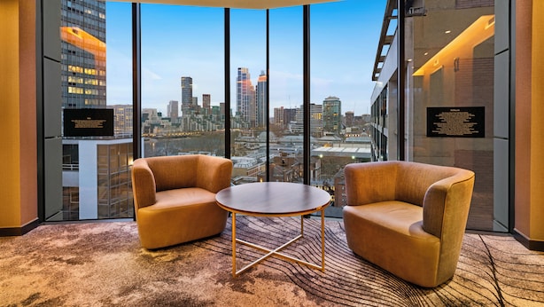 A corner room with two armchairs, a small round table, and a city view through large windows.