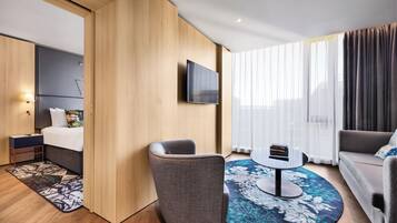 Ein modernes Hotelzimmer mit Bett, einer Sitzecke mit Tisch, einem an der Wand montierten Fernseher und großen Fenstern mit Vorhängen.