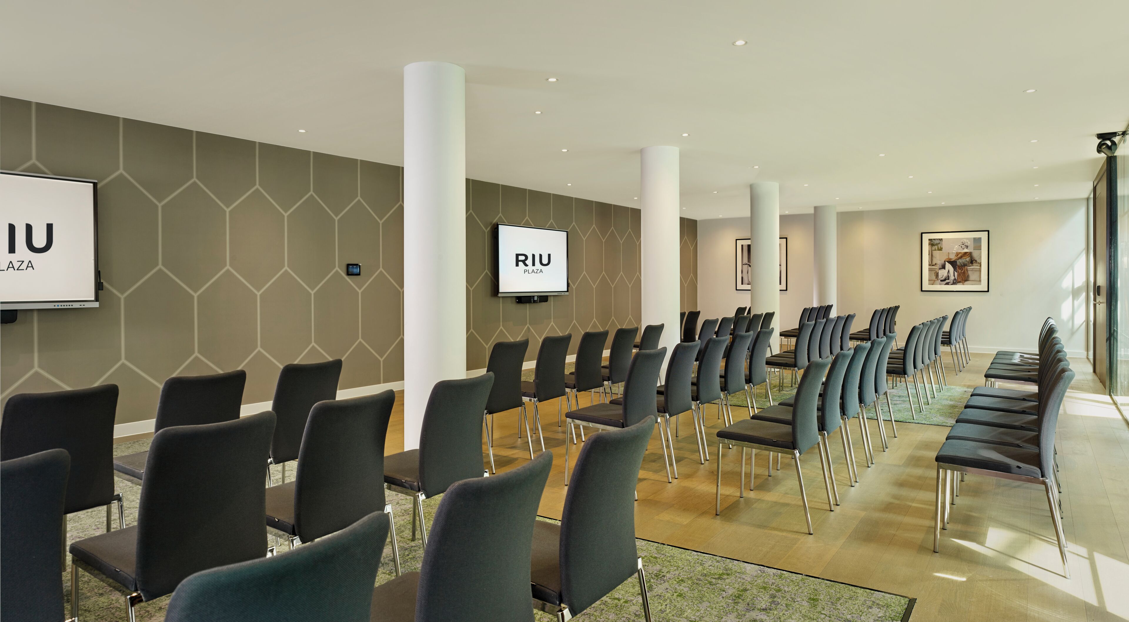 Una sala conferenze con file di sedie, un grande schermo e un cartello con la scritta 'RIU PLAZA'.