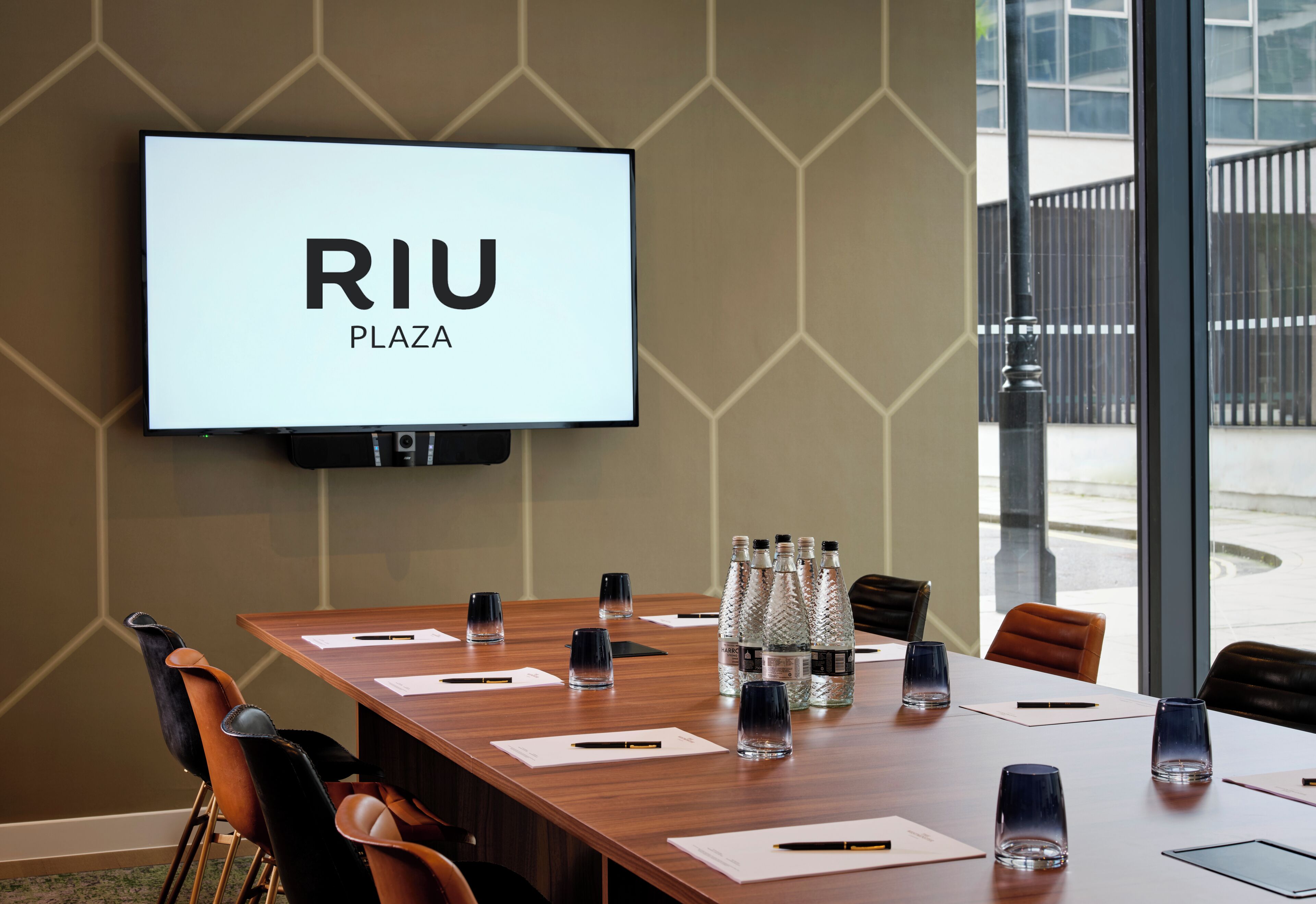 A conference room with a large screen displaying 'RIU PLAZA,' a long wooden table with chairs, water bottles, and documents.