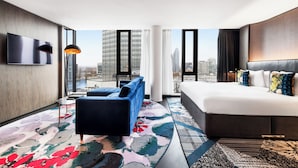 A modern hotel room with a large bed, a sofa, a TV mounted on the wall, and a city view.