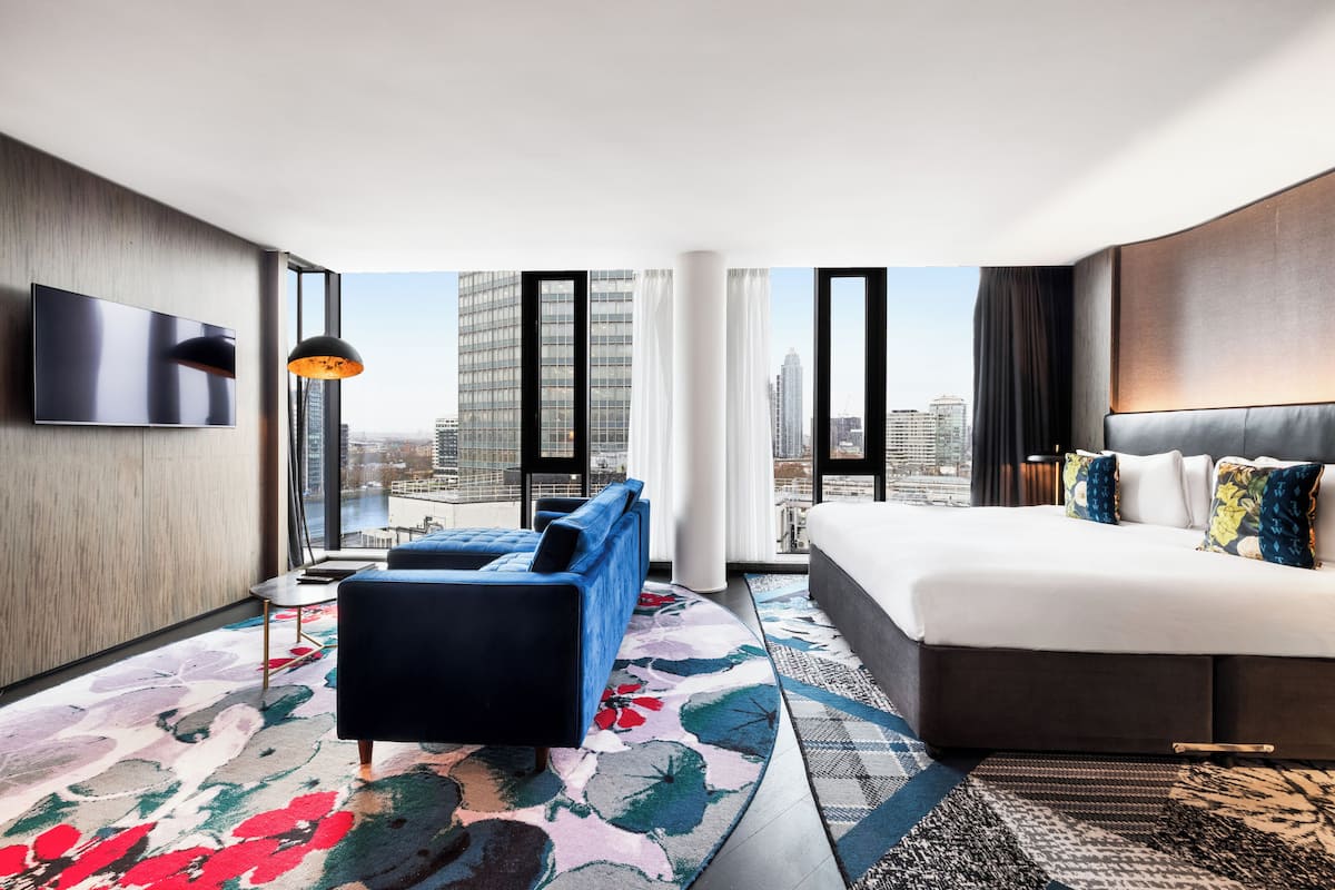A modern hotel room with a large bed, a sofa, a TV mounted on the wall, and a city view.