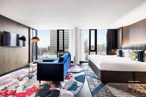 A modern hotel room with a large bed, a sofa, a TV mounted on the wall, and a city view.
