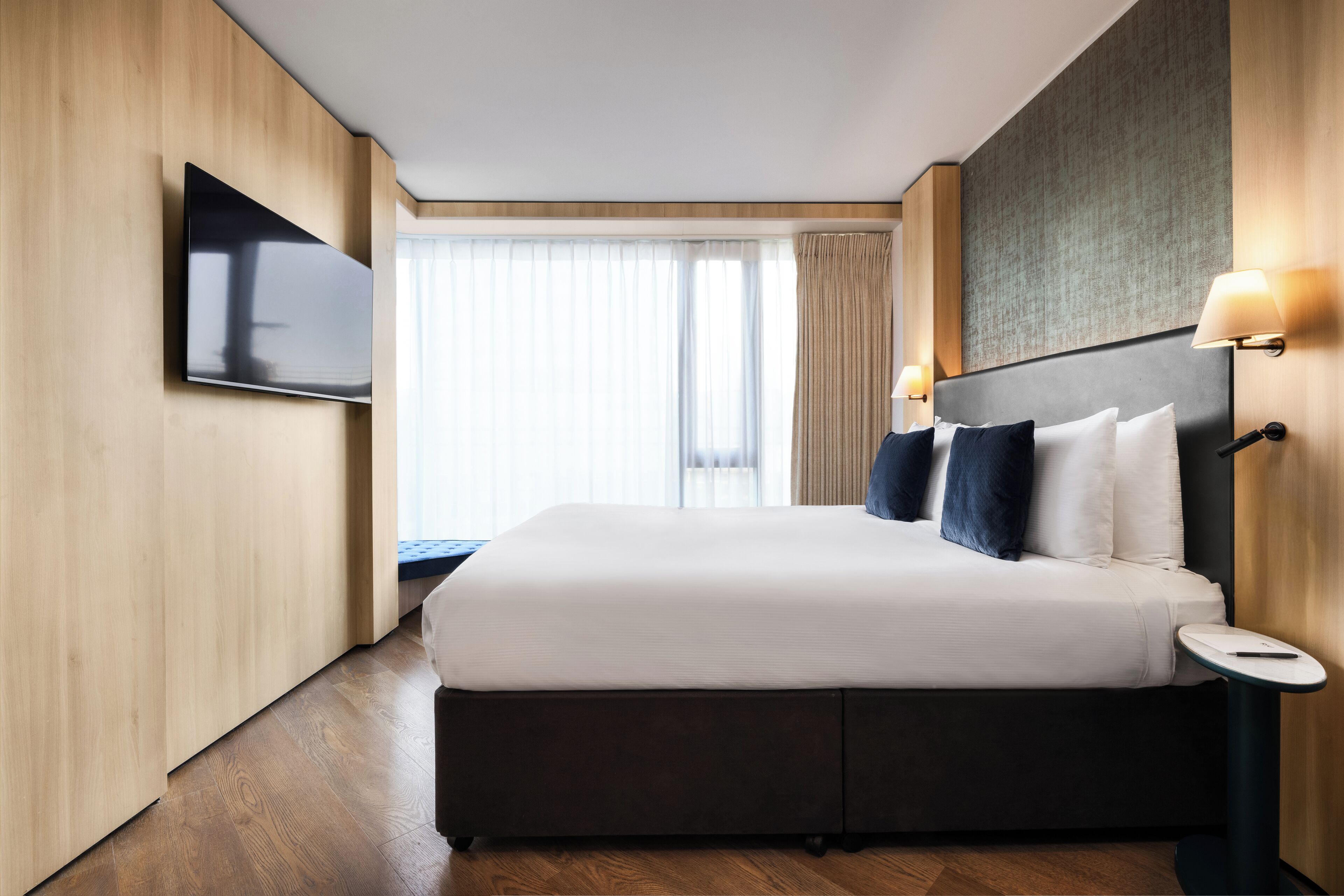 A modern hotel room with a large bed, a flat-screen TV mounted on the wall, and a small bedside table.