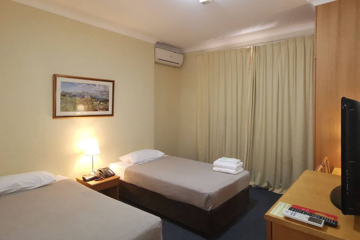 A hotel room with two beds, a desk with a lamp, a painting on the wall, and an air conditioning unit.