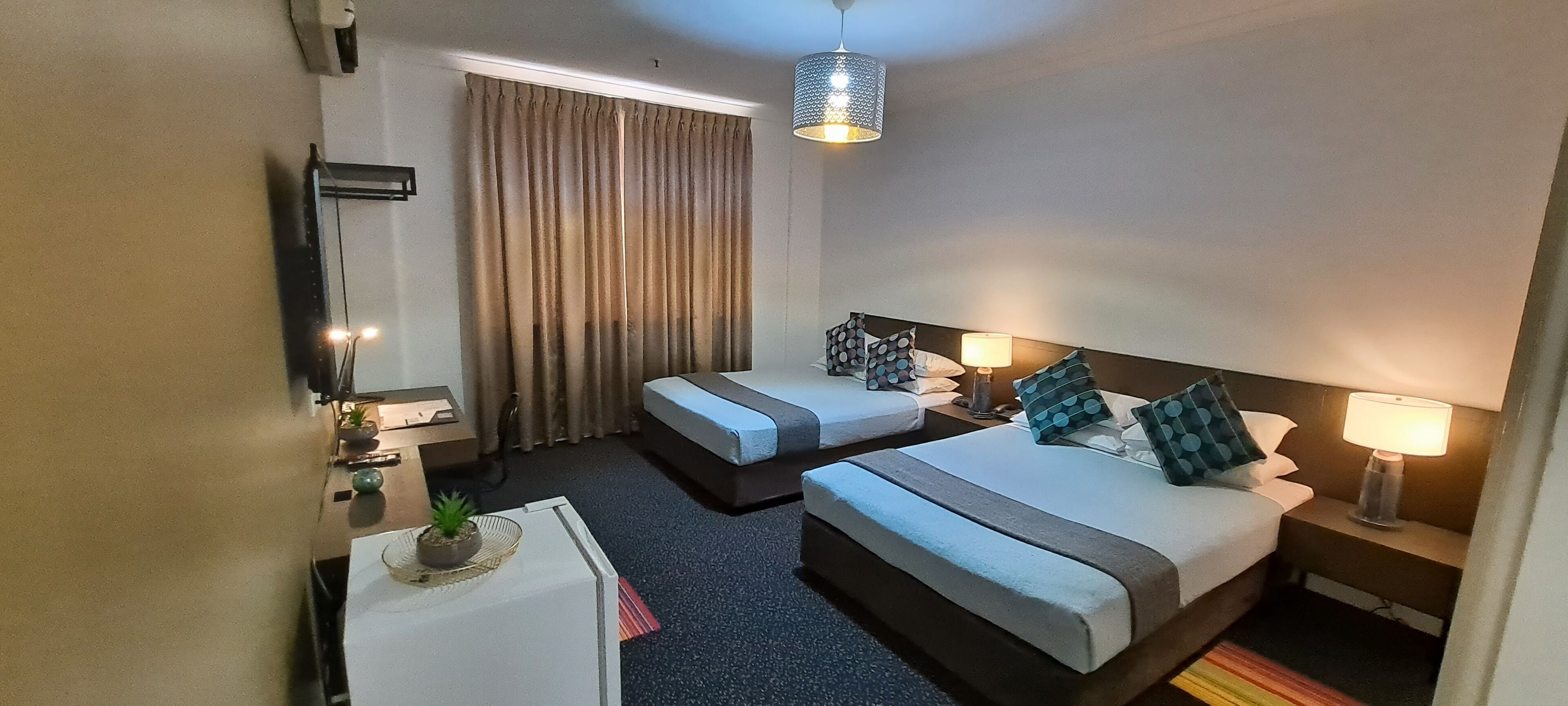 Une chambre d’hôtel avec deux lits, un petit bureau, une lampe, une plante en pot et un rideau.