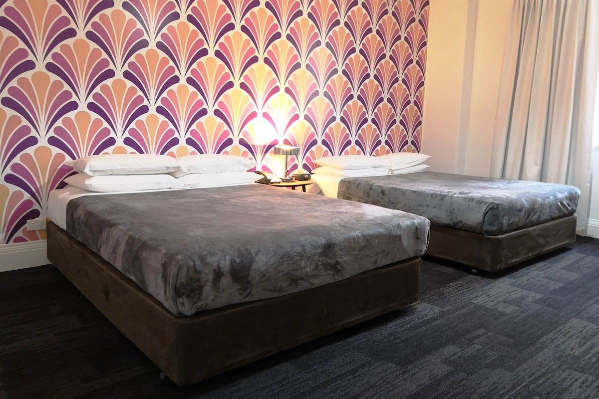 A hotel room with two beds, a patterned wallpaper, a lamp, and a window with curtains.